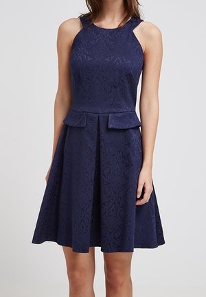 Kobieta w granatowej sukience bez rękawów z subtelnym kwiatowym wzorem i detalami peplum w talii, stojąca na jednolitym tle.