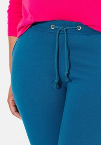 Teal collegebyxor med elastiskt midjeband, med dragsko med guldfärgat detaljer och fransar, tillverkade av mjukt tyg.