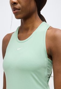Femme portant un haut sans manches Nike vert clair avec petit logo swoosh blanc, cheveux foncés tirés en arrière.
