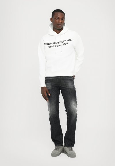 White hoodie with black text, paired with dark denim jeans featuring a distressed finish. Light gray sneakers complete the look.