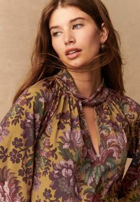 Blusa floreale realizzata in tessuto morbido, con scollatura a V profonda, colletto arricciato e pattern intricati in viola e verde su uno sfondo senape.