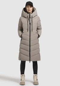 Beige gefütterter langer Mantel mit Kapuze, Frontreißverschluss und schwarzen Akzenten. Verfügt über ein gestepptes Design und Seitentaschen, getragen über schwarzen Leggings und beigen Stiefeln.