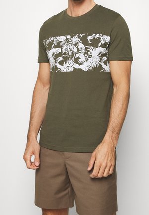 Mann trägt ein olivgrünes T-Shirt mit weißem tropischem Aufdruck auf der Brust und braune Shorts, steht vor einem weißen Hintergrund.