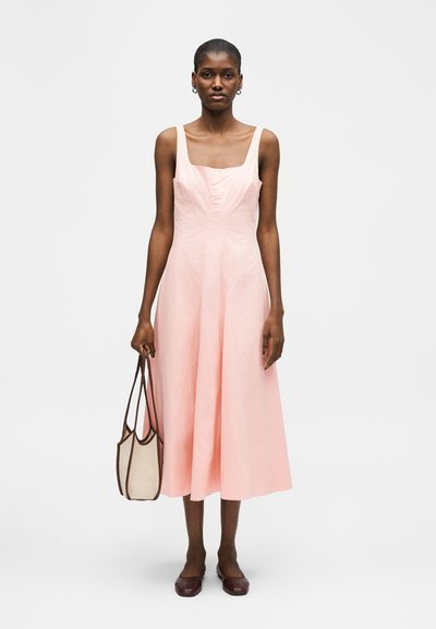 Frau steht und trägt ein ärmelloses hellrosa Midikleid, hält eine beige Tragetasche mit braunen Trägern und braune flache Schuhe.