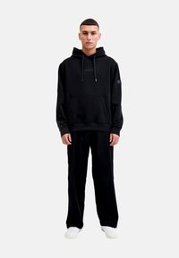 Schwarzer Hoodie mit Kordelzügen und Fronttasche, kombiniert mit schwarzen Hosen. Merkmale: Logo am Ärmel und gerippte Bündchen. Weicher Stoff, lockere Passform.