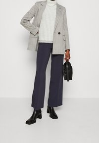Persona che indossa un blazer a righe, un dolcevita bianco a trama, pantaloni larghi blu navy, stivali neri con tacco, mentre tiene una piccola borsa nera di lato.