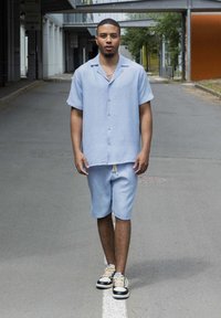 Chemise légère bleu clair à manches courtes avec une texture subtile, fermeture à boutons, et shorts assortis. Portée avec des baskets noires et une chaîne en argent.