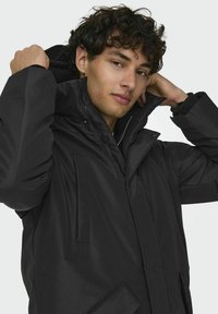Chaqueta aislante negra con cuello alto, cierres de cremallera y botón a presión, bolsillos laterales y textura suave. El modelo ajusta la capucha.