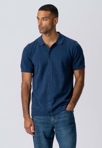 Marineblå kortærmet polo skjorte lavet af struktureret stof, med klassisk krave, knappelukning og ribbede manchetter, parret med blå jeans.