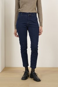 Sötétkék farmernadrág, slim fit szabással, gombos záródással és felhajtott szárakkal, fekete cipővel és világosbarna pulóverrel párosítva.