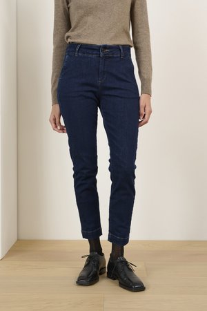 Tumed sinised denim püksid kitsase sobivusega, millel on nupuväline sulgemine ja volditud pahkluude osa, koos mustade kingade ja heleda pruunika sviitriga.