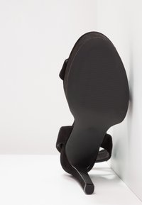Sandale noire à talon haut reposant sur son talon, montrant une semelle lisse et un talon fin sur fond blanc et mur d'angle.