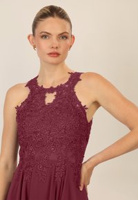 Burgundowa sukienka z misternym koronkowym detalem w kwiaty, bez rękawów, z wysokim dekoltem i wycięciem w kształcie łezki. Gładka tkanina układa się elegancko.