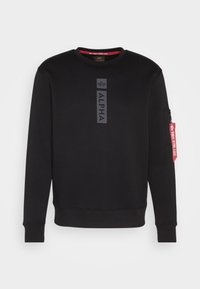 Sweat-shirt noir à manches longues avec le texte "ALPHA" et un logo sur la poitrine, poignets et ourlet côtelés, et une étiquette rouge "REMOVE BEFORE FLIGHT" sur la poche de la manche gauche.