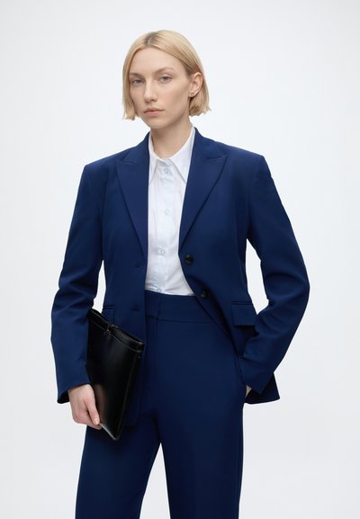 Femme en costume bleu marine et chemise blanche tenant un dossier en cuir noir, debout devant un fond clair uni.
