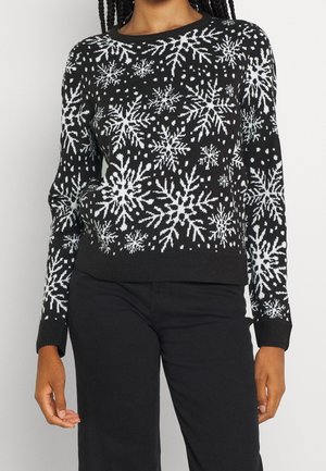 Femme portant un pull noir avec des motifs de flocons de neige blancs et un pantalon noir, se tenant devant un fond clair uni.