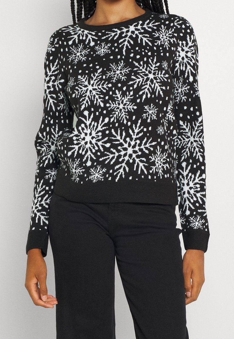 Femme portant un pull noir avec des motifs de flocons de neige blancs et un pantalon noir, se tenant devant un fond clair uni.
