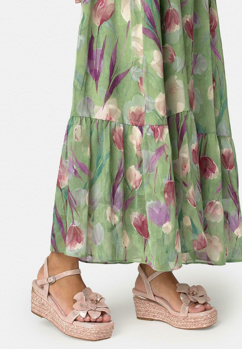 Pieds portant des sandales compensées roses à motifs floraux avec brides à la cheville sous une robe longue verte à fleurs.