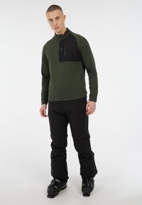 Groene trui met lange mouwen en een zwart borstpaneel, met ritskraag, in combinatie met zwarte broek en skischoenen. Gladde textuur en strak design.