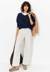 Pull en V en maille marine, pantalon large crème à imprimé floral bleu, veste beige enroulée sur le bras, et chaussures à enfiler beige.