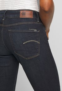 Tmavě modré džíny s úzkým střihem, s hnědým koženým štítkem na pase, kontrastním šitím a dvěma zadními kapsami.