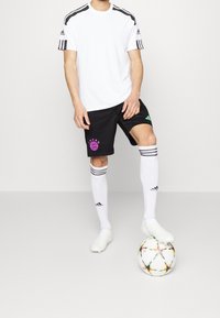 Maglietta sportiva bianca con strisce nere, pantaloni corti neri con un badge rosa, calze bianche con dettagli grigi e scarpe chiare, in piedi accanto a un pallone da calcio.