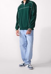 Grüne Fleecejacke mit halbem Reißverschluss und weißen Paspeln, kombiniert mit locker sitzenden hellblauen Jeans und dunkelblauen Sneakers mit grauen Akzenten.