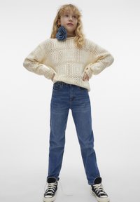 Knitad krämfärgad tröja med ett mönstrat design, kombinerad med blå jeans och svarta högskaftade sneakers. En blå blomma som accessoar bärs runt halsen.