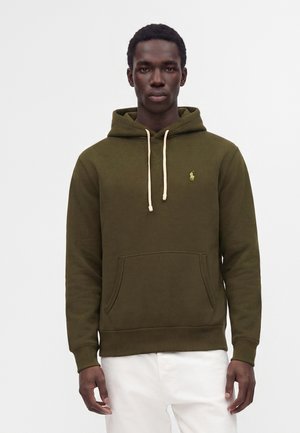 Homme portant un sweat à capuche vert olive avec des cordons blancs et un petit logo jaune sur la poitrine, associé à un pantalon blanc, debout face à l'avant.