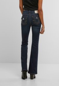 Jeans svasati di colore blu scuro realizzati in denim, con evidenti tasche posteriori dotate di ricami e hardware in argento. Texture liscia, taglio classico.