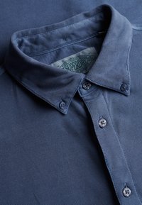 Knappet marineblå tekstureret poloshirt med knappet krave og synligt Superdry-mærke inde i halsudskæringen.