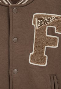 Hnedá univerzitná bunda s veľkým textúrovaným písmenom „F“ na náplasti a emblémom vlajočky „GO TEAM“ nad písmom na hrudi.