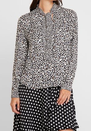 Femme portant un chemisier à manches longues à imprimé animalier avec un nœud à l'avant et une jupe noire à gros pois blancs.
