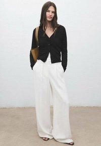 Black, long-sleeve cardigan with buttons, worn over a white top. Loose-fitting white trousers and a brown bag complete the outfit.