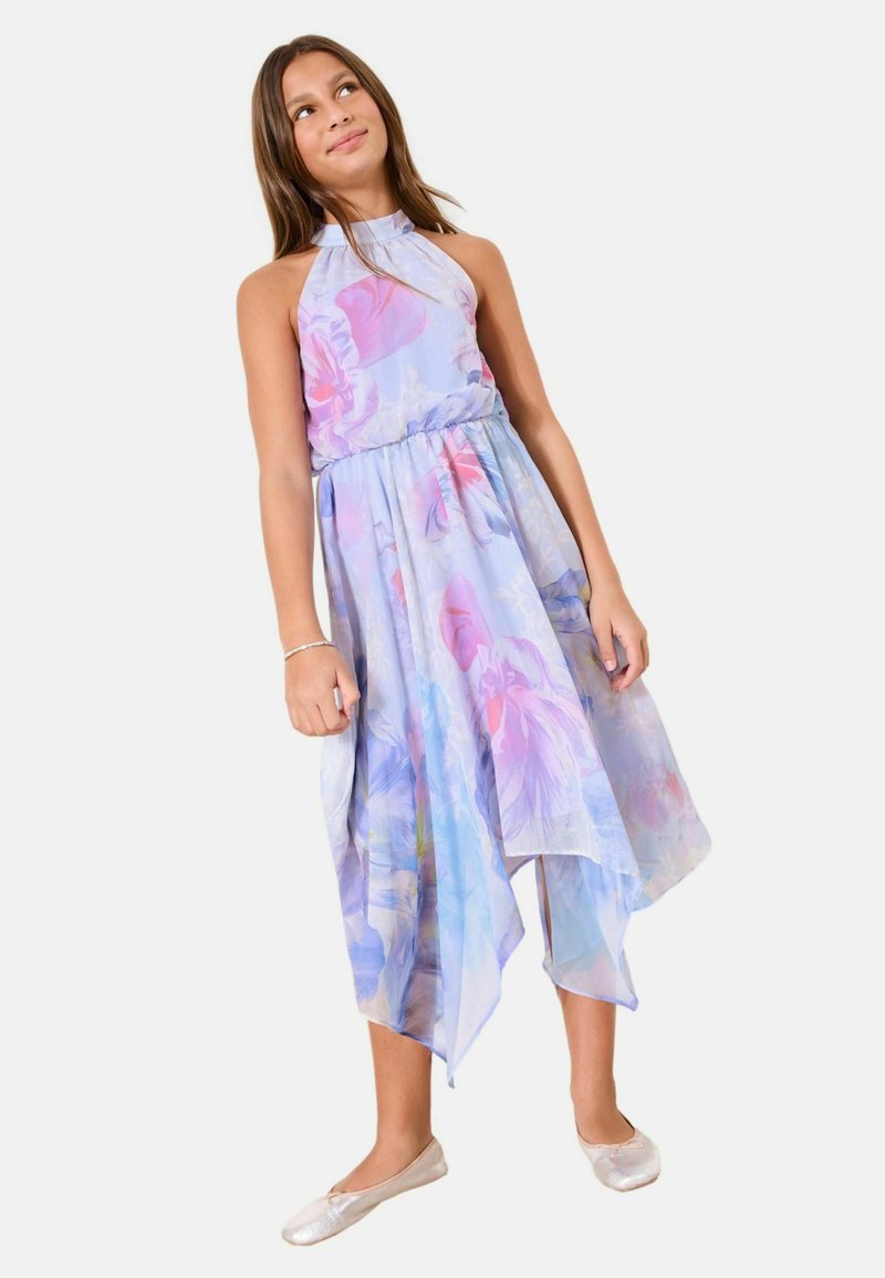 Niña vestida con un vestido sin mangas de flores en tonos pastel con un dobladillo asimétrico, de pie y mirando ligeramente hacia arriba y hacia un lado.