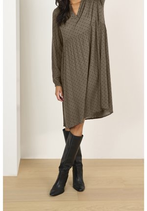 Woman wearing a long-sleeve olive green polka dot dress paired with black knee-high leather boots, standing on a wood floor.