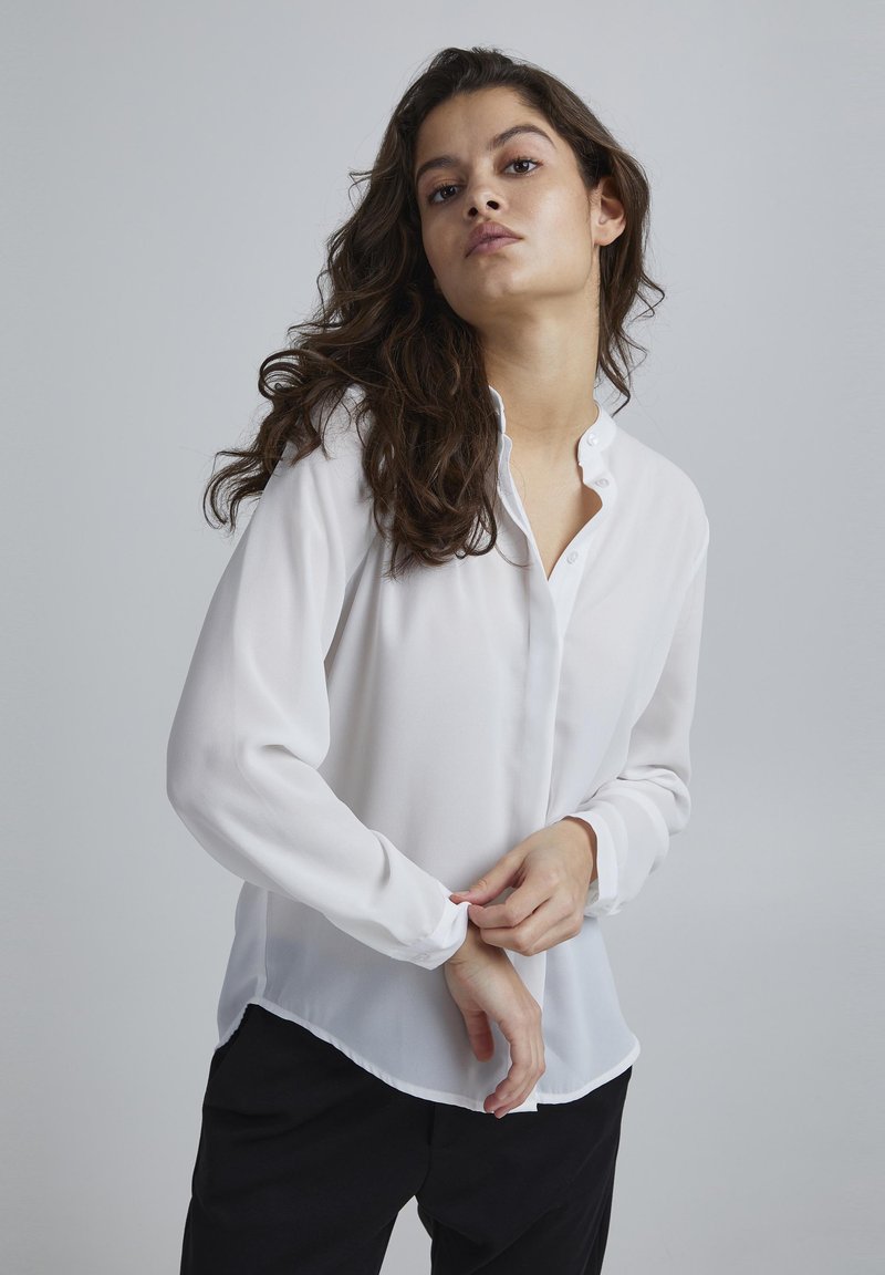 White, button-up blouse with long sleeves, featuring a soft, sheer texture and a rounded hem. Pairing with black pants completes the look.