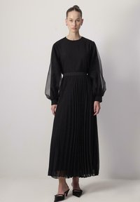 Zwarte geplooide maxi-jurk met lange doorschijnende mouwen, een ronde halslijn en een getailleerde taille. Geaccentueerd met een smalle tailleband voor extra definitie.