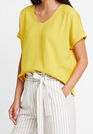 Vrouw draagt een gele V-hals blouse met korte mouwen, ingestopt in een witte broek met hoge taille, verticale strepen en een stoffen ceintuur die is vastgeknoopt.