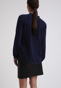 Blusa blu navy con maniche lunghe e polsini con bottoni, caratterizzata da una texture di tessuto liscia e un orlo arrotondato, abbinata a una gonna nera.
