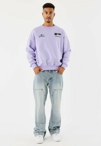 Hombre de pie de frente, vistiendo un sudadera lavanda, jeans de color azul claro con grandes bolsillos y zapatillas blancas, con las manos en los bolsillos.