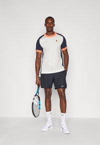 Ανδρική αθλητική ενδυμασία με ανοιχτό γκρι μπλούζα, ναυτικές μανσέτες και πορτοκαλί λεπτομέρειες, συνδυασμένη με μαύρες βερμούδες και λευκά αθλητικά παπούτσια. Κρατάει μια ρακέτα τένις.