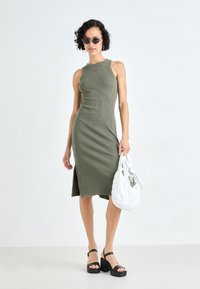 Olive green ribbed midi dress with a high neckline and side slit, paired with a textured white handbag and black platform sandals.