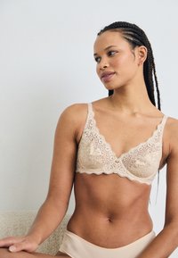 Lace bralette in nude, met klokvormige randen, verstelbare bandjes en bloemmotieven; zachte textuur, met een centrale strikdetail.