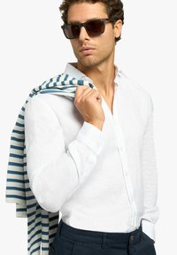 Witte linnen shirt met knopen, gecombineerd met marineblauwe broek. Een blauw-wit gestreepte gebreide trui over de schouder gedrapeerd. Bruine zonnebril.