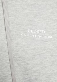 Sweat-shirt gris avec une texture douce. Il présente un texte blanc à l'avant "CLOSED" et "Creative Department". Comprend un cordon de serrage pour l'ajustement.