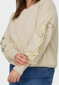 Kermanvärinen collegepaita, jossa on kultaiset kukkakuviot hihoissa, resorikahleet ja rento istuvuus. Pehmeä tekstuuri ja pyöreä pääntie.