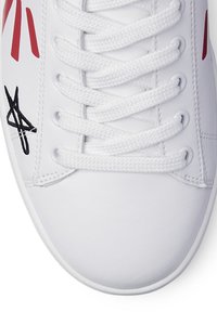 Baskets blanches avec des accents rouges, tige synthétique texturée, lacets blancs plats, et détail du logo noir sur le côté. Conception à bout rond.