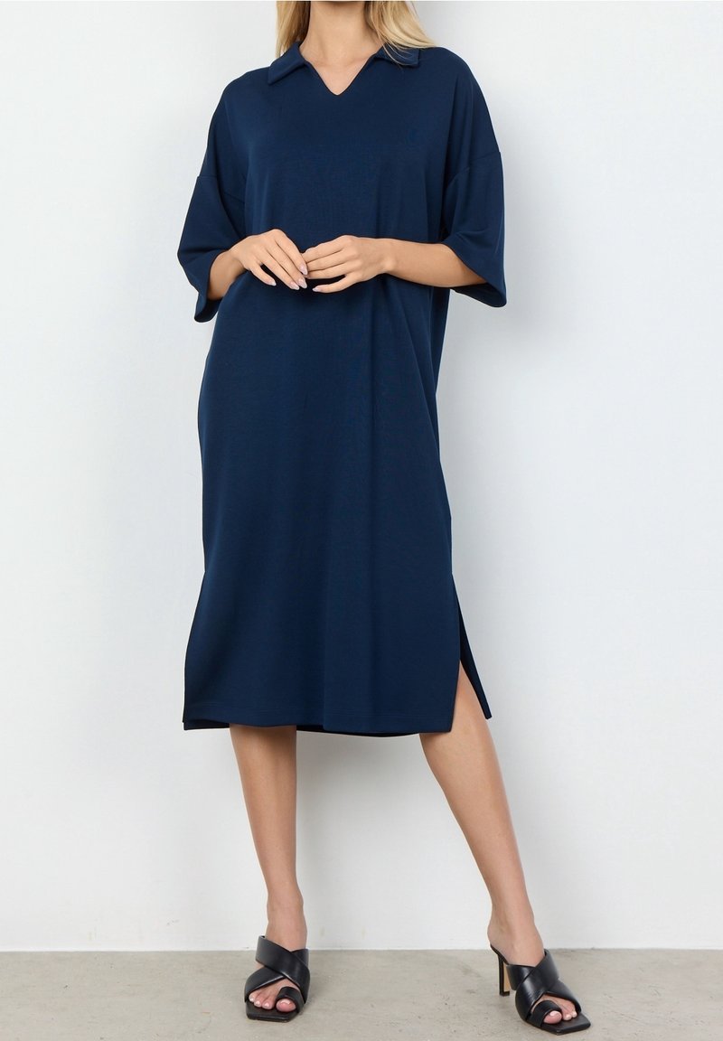 Femme portant une robe bleu marine ample, longueur genou, à manches courtes, et des sandales noires à talons, debout contre un mur blanc uni.