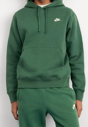Osoba nosząca zieloną bluzę z kapturem Nike i pasujące spodnie dresowe z białymi logo, stoi z rozluźnionymi rękami wzdłuż ciała.